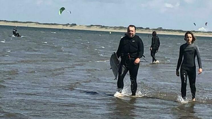 Marko en twee anderen in wetsuits, lopend door ondiep water, met kitesurfers op de achtergrond.